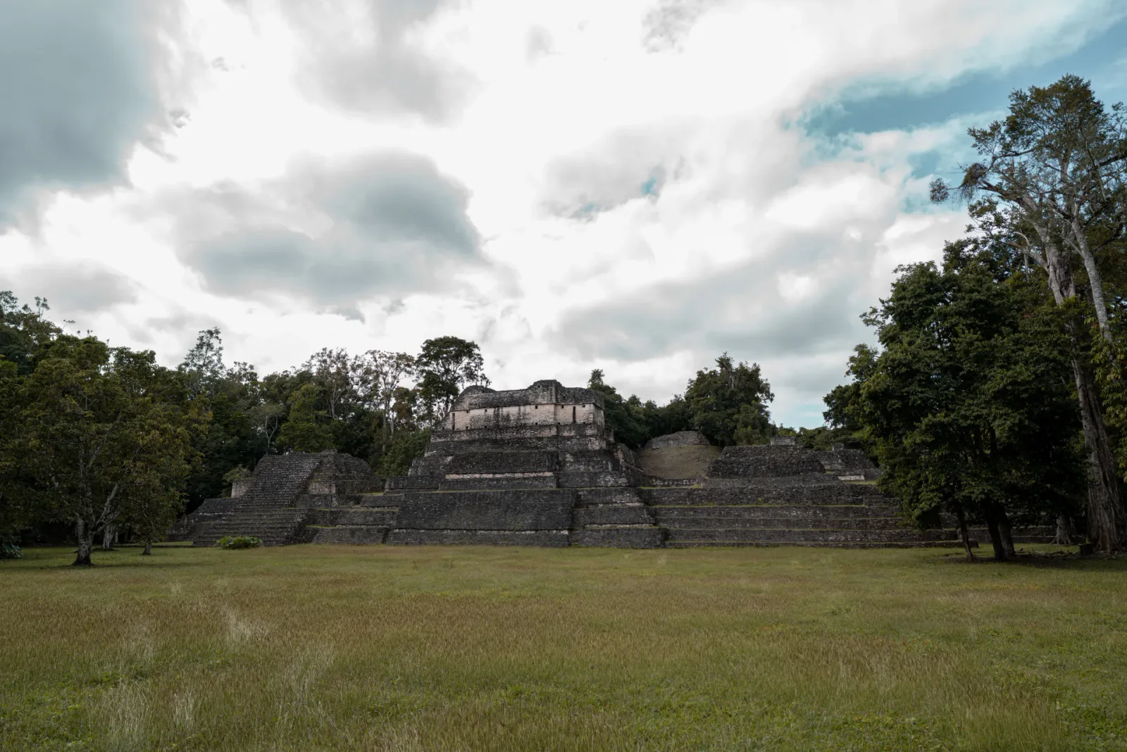 Ancient Maya buildings at Caracol surrounded by rainforest beneath a dramatic cloudy sky.